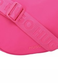 Borsa in nylon rosa con una ampia tracolla che presenta il testo "HUGO" in rilievo. Il design è liscio e arrotondato con una finitura opaca.