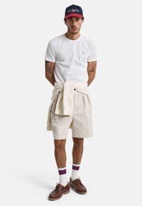 Hvid t-shirt, beige shorts, brune sko og multifarvede stribede sokker. Bærer en marineblå og rød kasket. Sweater bundet om livet.