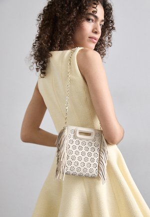 Mujer con cabello rizado que lleva un vestido amarillo sin mangas, portando un bolso beige con recortes florales, flecos y una cadena dorada como correa.