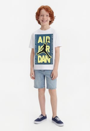 Ragazzo sorridente con i capelli rossi in piedi, indossa una maglietta bianca con grafica Air Jordan, pantaloncini di jeans azzurri e scarpe da ginnastica blu navy con lacci bianchi.