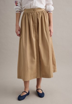 Donna che indossa una gonna midi beige a vita alta con pieghe, camicetta bianca con polsini ricamati colorati e scarpe Mary Jane blu scuro.