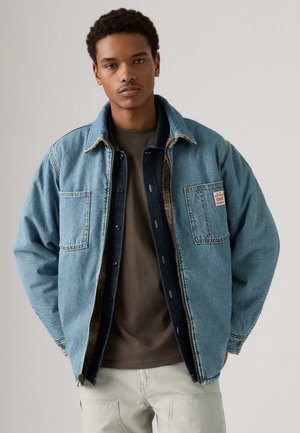 Denim jacket with a light blue exterior, two pockets, and dark denim lining; worn over a dark gray shirt and beige pants.