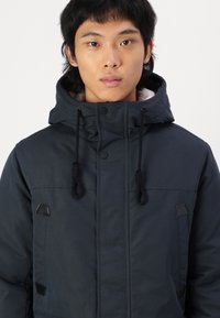 Chaqueta acolchada azul marino con capucha, cordones ajustables, detalles en negro, bolsillos delanteros y un forro interior suave.