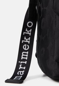 Musta reppu, jossa on kohokuvioitu kukkakuosi. Varustettu tukevalla hihnalla, jossa lukee "marimekko" valkoisella, teksturoidulla kankaalla, ja se on pyöreän muotoinen.