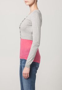 Grå virkade cardigan med knappar och långa ärmar, lagd över en rosa skjorta. Bärs med blå denimjeans som framhäver en åtsittande design.