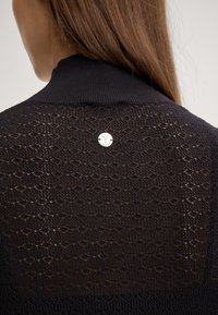 Maglione nero lavorato a maglia con un motivo testurizzato, colletto alto e un accento logo argentato sulla parte posteriore. Design a trama aperta per una migliore traspirabilità.