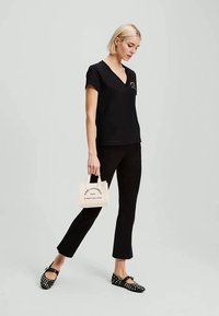 T-shirt preta de decote em V, calças pretas ajustadas e sapatos pretos com tachas. A segurar uma pequena mala beje com logótipo e texto impressos.