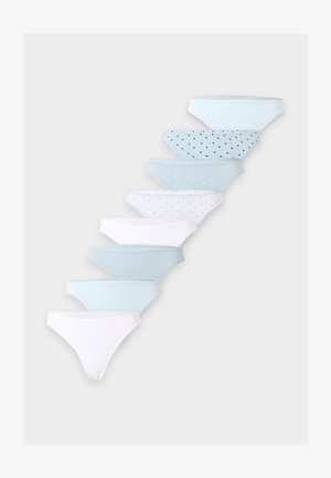 Jeune femme portant un soutien-gorge beige et une culotte bleu clair à pois, debout avec les bras derrière le dos sur un fond blanc uni.