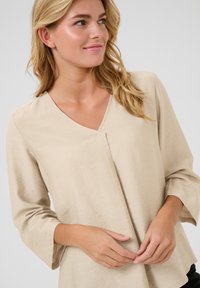 Blouse beige à col en V en tissu léger, avec des manches trois-quarts et une coupe décontractée, présentant une texture subtile. Détails de surface lisse.