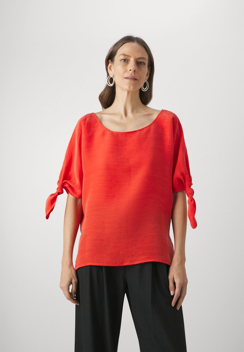 Red, short-sleeved top with a wide neckline and tied sleeves. Made of lightweight fabric with a subtle texture. Paired with black trousers.