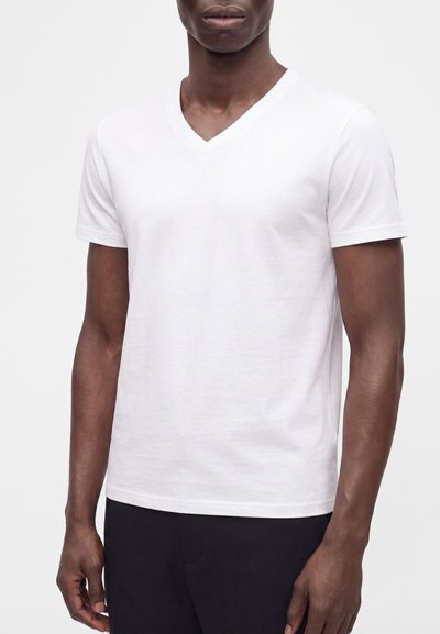 Άνδρας που φοράει απλό λευκό κοντομάνικο T-shirt με V-λαιμόκοψη και μαύρο παντελόνι, στέκεται μπροστά σε ένα απλό φωτεινό φόντο.