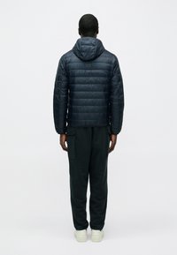 Sötétkék pufi dzseki kapucnival, steppelt panelokkal és rövidített kialakítással. Sötétzöld melegítőnadrággal és fehér sportcipővel párosítva.