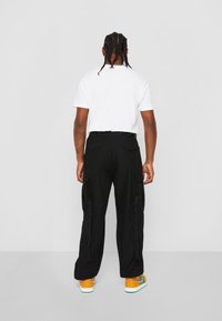 Pantalones cargo negros con bolsillos laterales, textura de la tela visible. Combinados con una camiseta blanca y zapatillas de colores, vistos desde atrás.