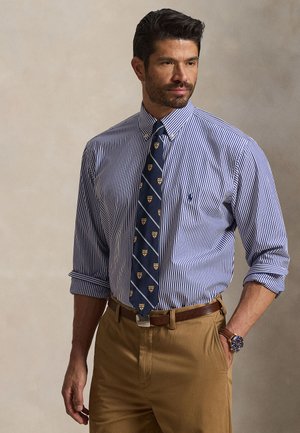 Homme aux cheveux foncés et à la barbe porte une chemise à rayures bleues boutonnée, une cravate bleu marine à motifs, un pantalon beige, une ceinture marron et une montre-bracelet, debout avec une main dans la poche.