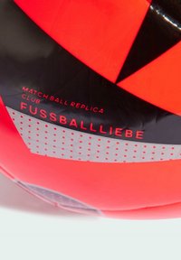 Palla da calcio rossa e nera con superficie sintetica liscia, caratterizzata da un design geometrico e testo: "REPLICA DELLA PALLA DA MATCH CLUB FUSSBALLLIEBE."
