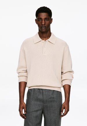 Beiger Strick-Polo-Pullover mit Rippstruktur, Drei-Knopfleiste und langärmeligen, mit Bündchen versehenen Ärmeln, kombiniert mit grauen Nadelstreifenhosen.