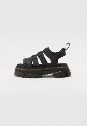 Black platform sandal made of synthetic material, featuring three adjustable straps, a textured sole, and contrasting yellow stitching.