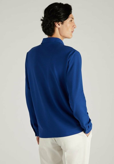 Polo à manches longues en bleu royal, fabriqué en tissu lisse, doté d'un col classique et de poignets boutonnés, avec une coupe moderne.