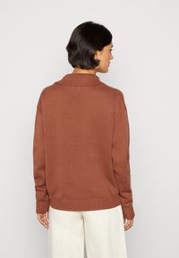 Brun strikket sweater med krave, der har et struktureret mønster og ribbet kant. Bæres med lyse bukser. Vist i ryggen.