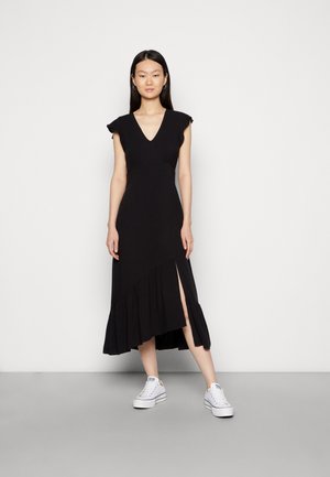 Vestido midi preto com mangas curtas, decote em V, corpete ajustado e bainha assimétrica com babados. Apresenta um fenda lateral e é usado com ténis brancos.
