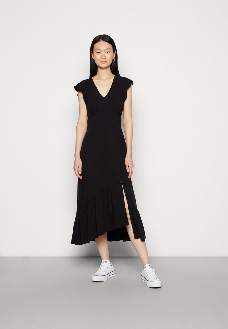 Vestido midi preto com mangas curtas, decote em V, corpete ajustado e bainha assimétrica com babados. Apresenta um fenda lateral e é usado com ténis brancos.