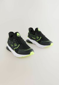 Black athletic shoes with green accents, mesh upper, cushioned white sole, and textured detailing along the sides. Adjustable laces.