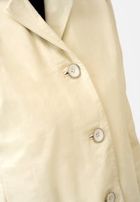 Krämfärgad läderblazer med en mjuk yta, med snedskurna slag och tre vita knappar fram.