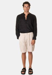 Camicia nera in lino a maniche lunghe con pattina, abbinata a pantaloni cargo beige con tasche laterali e una vestibilità rilassata, indossata con sandali neri.