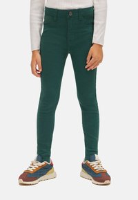 Pantalones de mezclilla verdes con un diseño ajustado, que cuentan con un cierre de botón y dos bolsillos frontales, combinados con zapatillas deportivas multicolores.