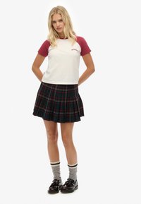 Witte t-shirt met wijnrode raglan mouwen en logo, gecombineerd met een geplooide zwart-groen geruite rok, grijs gestreepte sokken en zwarte schoenen.