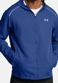 Chaqueta azul con cremallera y capucha, de textura lisa, con franjas de acento blancas en los hombros y un logo de Under Armour en el pecho.