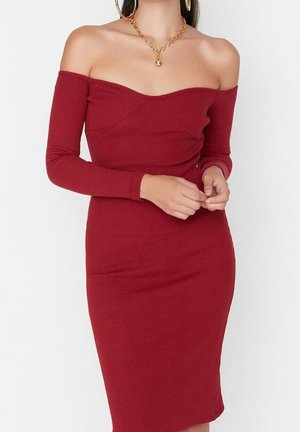 Kobieta w dopasowanej, czerwona sukience z długim rękawem odkrywająca ramiona, nosząca duże złote koła kolczyków i złoty łańcuszek z okrągłym wisiorkiem.