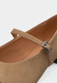 Chaussure Mary Jane en daim beige avec bride ajustable et boucle argentée, dotée d'un petit talon carré et d'une doublure intérieure lisse couleur cuir tan.