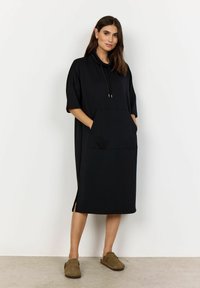 Zwarte oversized jurk met een capuchon, korte mouwen, een voorzak en zijsplitten. Gemaakt van een zacht materiaal met een gladde textuur.