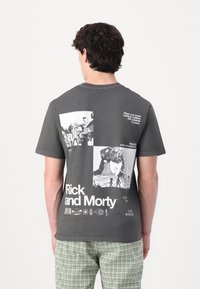 Sötétszürke pamut póló fehér grafikai nyomtatásokkal, beleértve a "Rick and Morty" feliratot és képregény stílusú illusztrációkat a hátsó részén.