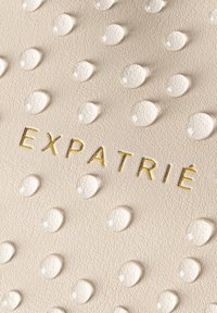 Pelle di colore crema con una texture a gocce rialzate. Scritta in oro "EXPATRIÉ" impressa al centro. Design liscio ed elegante con una leggera lucentezza.