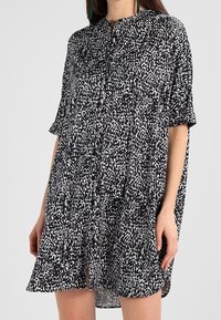 Mustavalkoinen lyhythihainen mekko löysällä istuvuudella. Siinä on pistekuvio ja nappisuljenta kauluksessa, ja se on valmistettu kevyestä kankaasta.