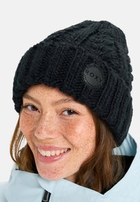 Black knitted beanie featuring a ribbed design and a prominent round black patch with the brand name "ROXY." Soft texture, warm material.