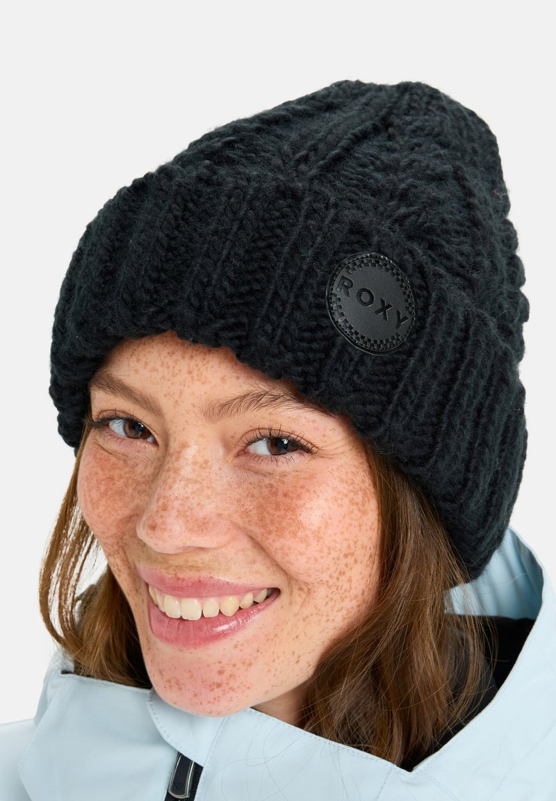 Black knitted beanie featuring a ribbed design and a prominent round black patch with the brand name "ROXY." Soft texture, warm material.