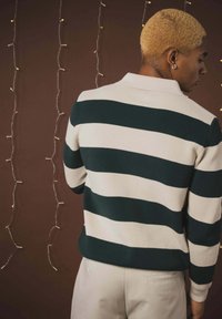 Pull en maille présentant de larges rayures horizontales vertes et blanches, une texture côtelée et un col à revers. Un pantalon de couleur claire est porté.