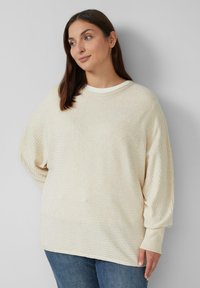 Kremowy, żeberkowy sweter z luźnym krojem, okrągłym dekoltem i długimi, lekko bufkami rękawami. W zestawie z dżinsami.
