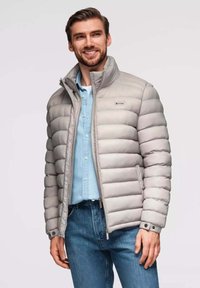 Helle graue Steppjacke mit horizontalem Quiltmuster, hohem Kragen, zwei-Wege-Reißverschluss und Druckknopfakzenten, kombiniert mit einem blauen Hemd und Jeans.