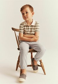 Gestreepte kortemouw polo in beige, bruin en groen, gecombineerd met grijze elastische broek en grijze instap schoenen, zittend op een houten stoel.