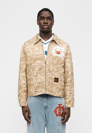 Νέος άνδρας φορώντας μπεζ ζακέτα με φερμουάρ και σχέδια, με λευκά και κόκκινα αφηρημένα μπαλώματα, ανοιχτόχρωμα μπλε τζιν με κόκκινο λεπτομερές μπαλώμα, στέκεται ακίνητος.