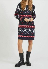 Robe pull de Noël bleu marine avec motifs de rennes et flocons de neige, accents rouges, manches longues et poignets côtelés ; associée à des bottes noires à semelles épaisses.