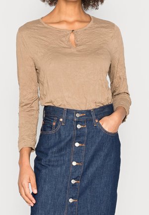 Gekräuseltes beiges Langarmhemd mit einem runden Halsausschnitt und einem Schlüsselloch. Kombiniert mit einem dunkelblauen Jeansrock mit Knöpfen vorne und Taschen.