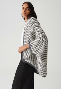 Femme portant un cardigan tricoté gris et blanc, ample, avec des manches larges, par-dessus un top blanc et un pantalon noir, se tenant devant un fond uni.