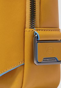 Angolo di una borsa sintetica gialla con finitura testurizzata, zip nera, cuciture blu e un accento in metallo con dettaglio del logo.