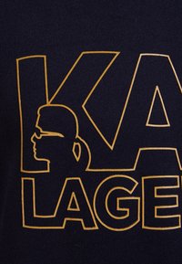 Granatowy materiał z dużymi złotymi literami napisanymi "KARL LAGERFELD", z sylwetką postaci w okularach przeciwsłonecznych.