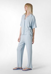 Lichtblauwe, kortlopende overhemd met knopen, gecombineerd met bijpassende losse broek. Soepele stof met een subtiele glans. Blauwe slippers maken de outfit compleet.